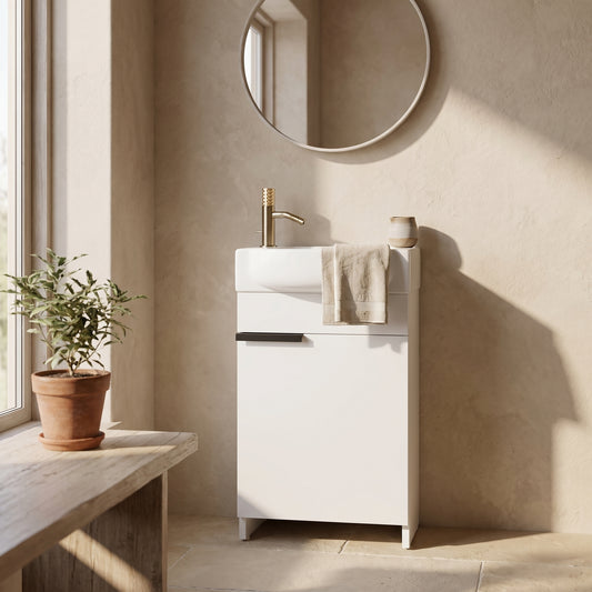 A minimalist bathroom vanity with a white finish, gold faucet, and a circular mirror above it. A potted plant sits on a wooden bench to the side.