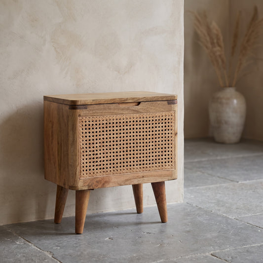 A wooden stool with a woven cane storage compartment, oak color, in a minimalist setting with a vase of reeds in the background