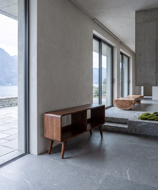 A dark wood shelf with a curved design, standing in a minimalist room with large windows looking out to a mountain and water backdrop.