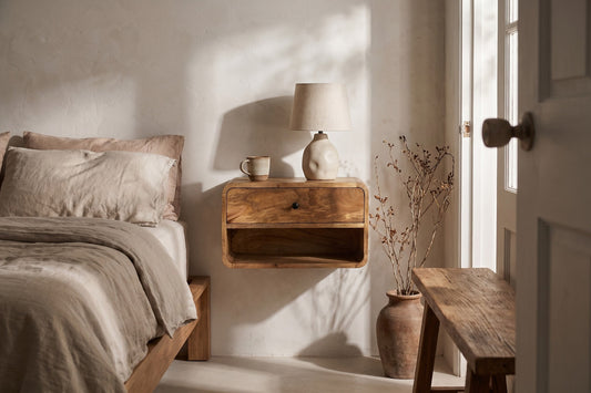 Solid wood bedside table with open shelf and drawer; positioned next to a bed with a beige linens and a lamp on top; light streams through a door with a wooden handle, creating a soft shadow on the wall.