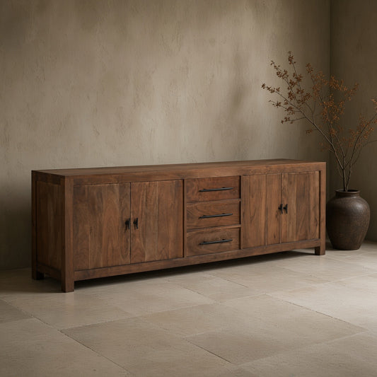 A wooden media console with four drawers and two cabinet doors, finished in brown, against a textured wall in a minimalist room with a vase of branches on the side.