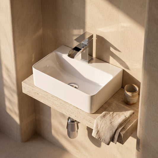 A wall-mounted, rectangular white ceramic bathroom sink with a modern chrome faucet. The sink is set against a beige wall with a textured surface and sits on a light-colored stone ledge. A cream-colored towel and a beige mug are placed nearby on the ledge.