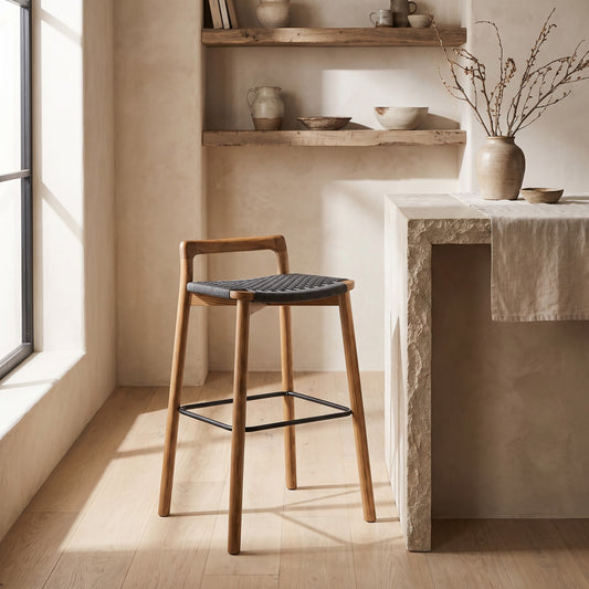 A modern minimalist wood barstool with a black seat and backrest, placed by a light-colored stone table in a room with wooden floors and wall-mounted shelves.
