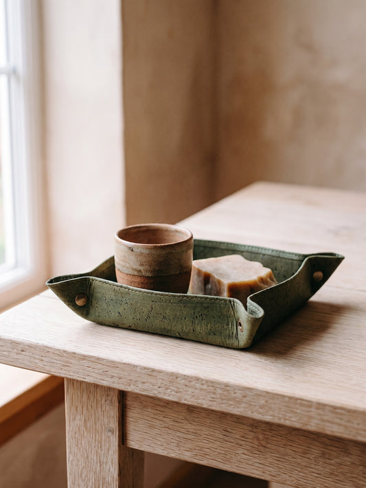 A small, green, speckled catch-all tray holds a ceramic cup and what appears to be a block of soap on a light wooden table in a minimal indoor setting.