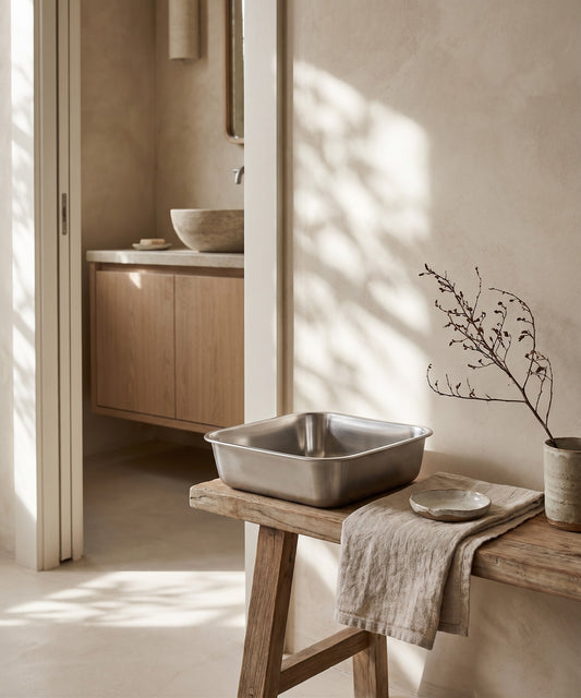 A square stainless steel bowl on a wooden table in a minimalist setting.