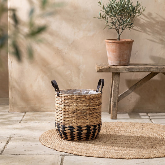 A two-tone basket planter with leather handles on a jute mat, background with a terracotta pot and wooden bench.