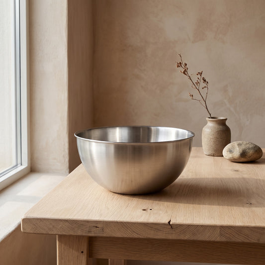 A stainless steel mixing bowl sits on a wooden table in front of a light-colored wall and window. A vase with a branch and a rock are also on the table. Smooth finish. Neutral tones.