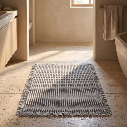 A textured grey bath rug with tassels on the edges, placed on terracotta tiles in a light and airy bathroom setting.