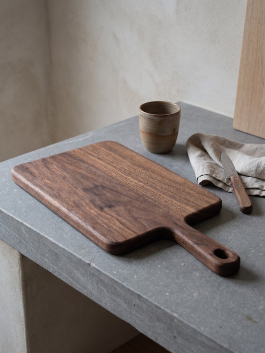 A wooden cutting board with a handle, placed on a concrete surface near a cup and a knife, against a neutral backdrop.