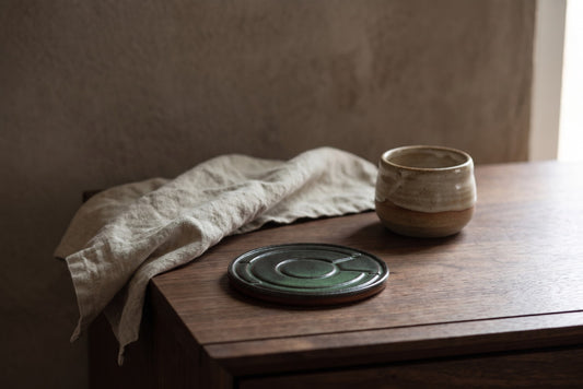 Green ceramic drink coaster with brown ring, placed on wooden table with tea cup and linen cloth in minimalist setting.