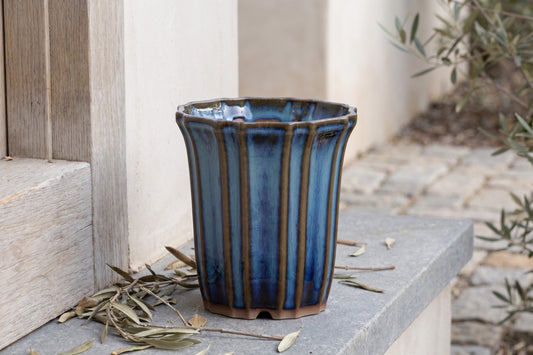 Rounded, ribbed, ceramic pot with a deep blue glaze, placed outdoors on a stone base near an olive tree and wooden door.