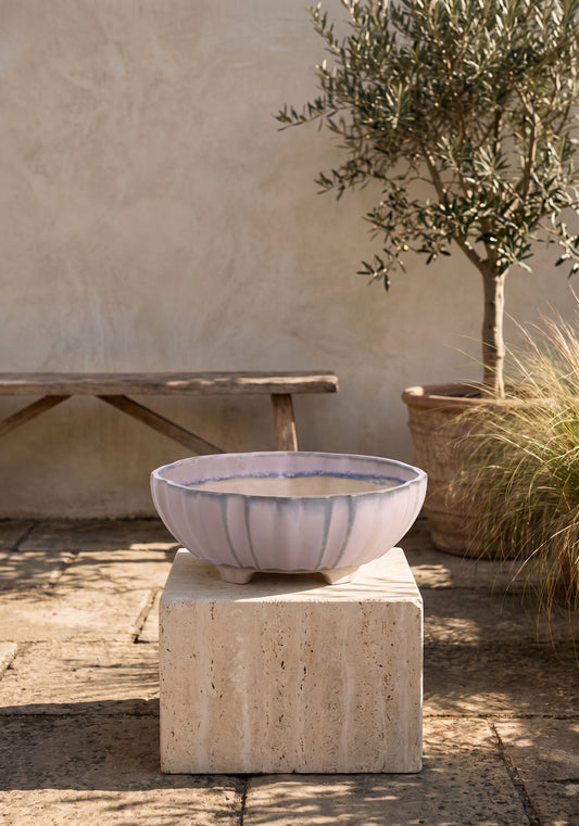 A round ceramic bonsai pot with blue and white stripes sits on a stone pedestal outdoors with a tree and wall in the background.
