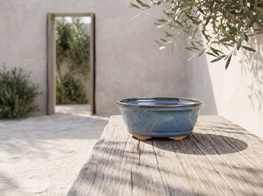 Oval ceramic bonsai pot 5.5 inch Peacock, blue glaze on a wooden table in front of a rustic wall and a tree branch with leaves.