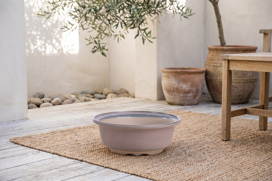 Round ceramic bonsai pot with ocean glaze placed on a woven mat on a wooden deck, with an olive tree and other planters in the background.