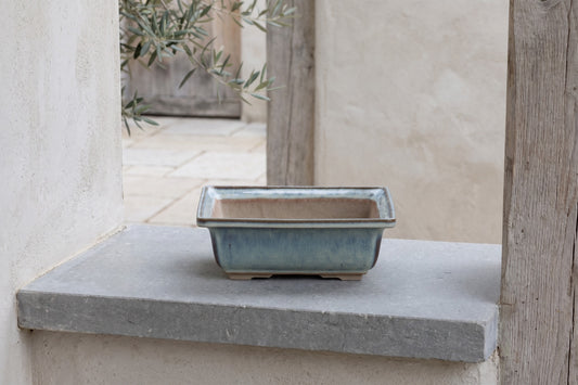 Square ceramic bonsai pot with a blue glaze, 7 inch Glacier, positioned on a wall shelf in a natural setting with stone and wood visible.