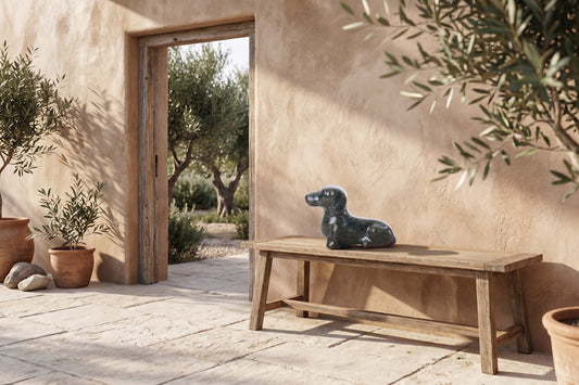 A ceramic dog figurine resembling a Dachshund with seaweed-like texture sits on a wooden bench in an outdoor setting with clay pots and greenery around.