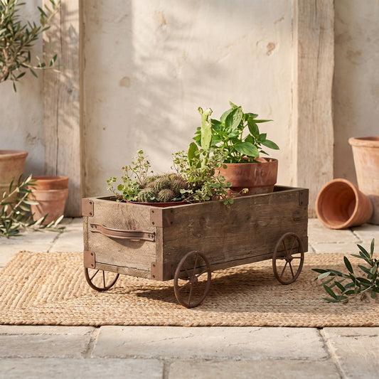 Wooden wagon planter with metal wheels and handle, filled with plants, on a woven mat.