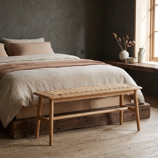 A simple, light brown Woven Design Bench placed in front of a bed with neutral bedding, wooden floor, and a window with natural light.