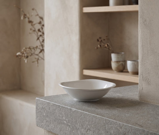 A white ceramic bowl with rounded corners sits on a stone shelf in a minimalist, modern setting with pale walls and shelves holding other pottery.