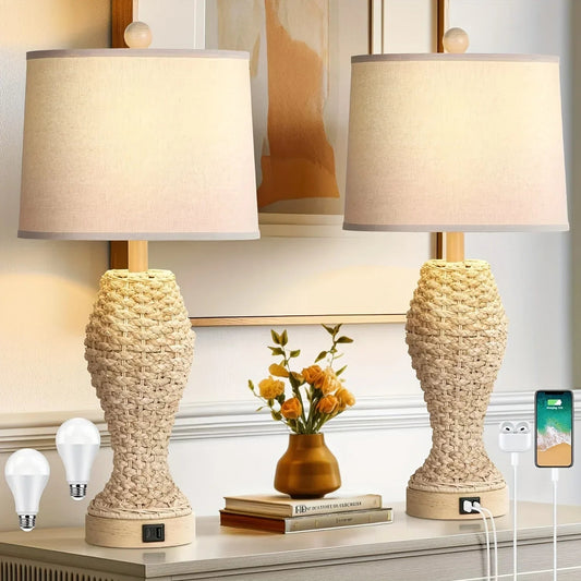 Two woven seagrass table lamps with cream fabric shades, plug, two lightbulbs, vase with yellow flowers, stack of books, phone on charger, placed on a dresser in a living room setting.
