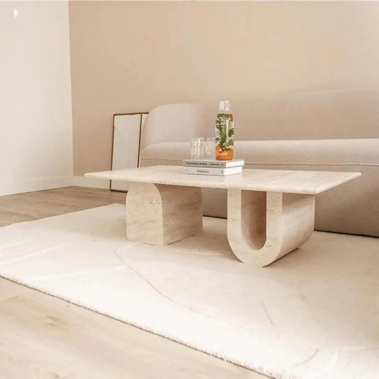 A U-shaped travertine coffee table in a minimalist setting with a vase and books on top, placed in front of a white sofa on a textured white rug.